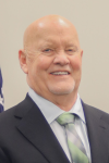 Bald white man in dark suit with blue shirt and tie in front of light background